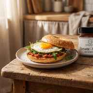 Ein aufgeklappter Sesambagel mit rotem Aufstrich, Rucola und Spiegelei, daneben ein Glas Tomate Walnuss