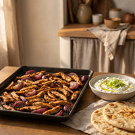 Knusprige Hähnchen-Gyros-Streifen mit roten Zwiebeln auf einem Backblech, daneben eine Schale Tzatziki