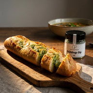 Ein aufgeschnittenes, goldbraun gebackenes Kräuterbaguette mit grüner Butter, daneben ein Glas Frühlings-Dip