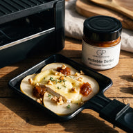 Ein Raclette-Pfännchen mit geschmolzenem Ziegenkäse, Birnenscheiben und Walnüssen, daneben ein Glas Verliebte Dattel