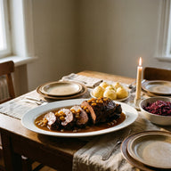 Scheiben von dunkel geschmortem Sauerbraten in glänzender brauner Sauce mit Rosinen, daneben Kartoffelklöße und Rotkohl