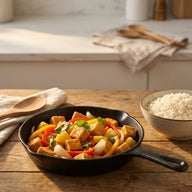 Eine Pfanne mit knusprigen Tofuwürfeln, roter und gelber Paprika und Ananas in glänzender Sauce, daneben Reis