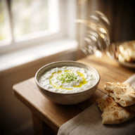Eine Schale cremiges Tzatziki mit Gurkenraspeln, Olivenöl und Fladenbrot daneben