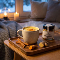 Eine Tasse Golden Milk mit Zimt und Ingwer auf einem Tablett in einer Sofaecke am Winterabend, daneben Golden Milk von Genusspower