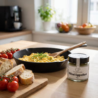 Rührei mit Dill in einer Gusspfanne, Brot und Tomaten am Küchentisch im Morgenlicht, daneben Rührei Eggcellent von Genusspower