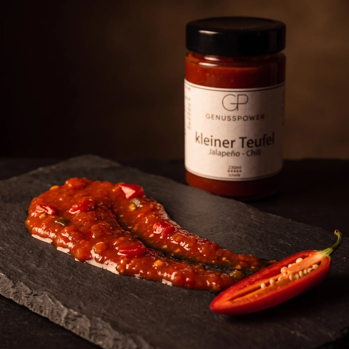 Stückige Sauce kleiner Teufel als Spur auf dunklem Schiefer, daneben eine halbierte rote Jalapeño, dahinter das Glas