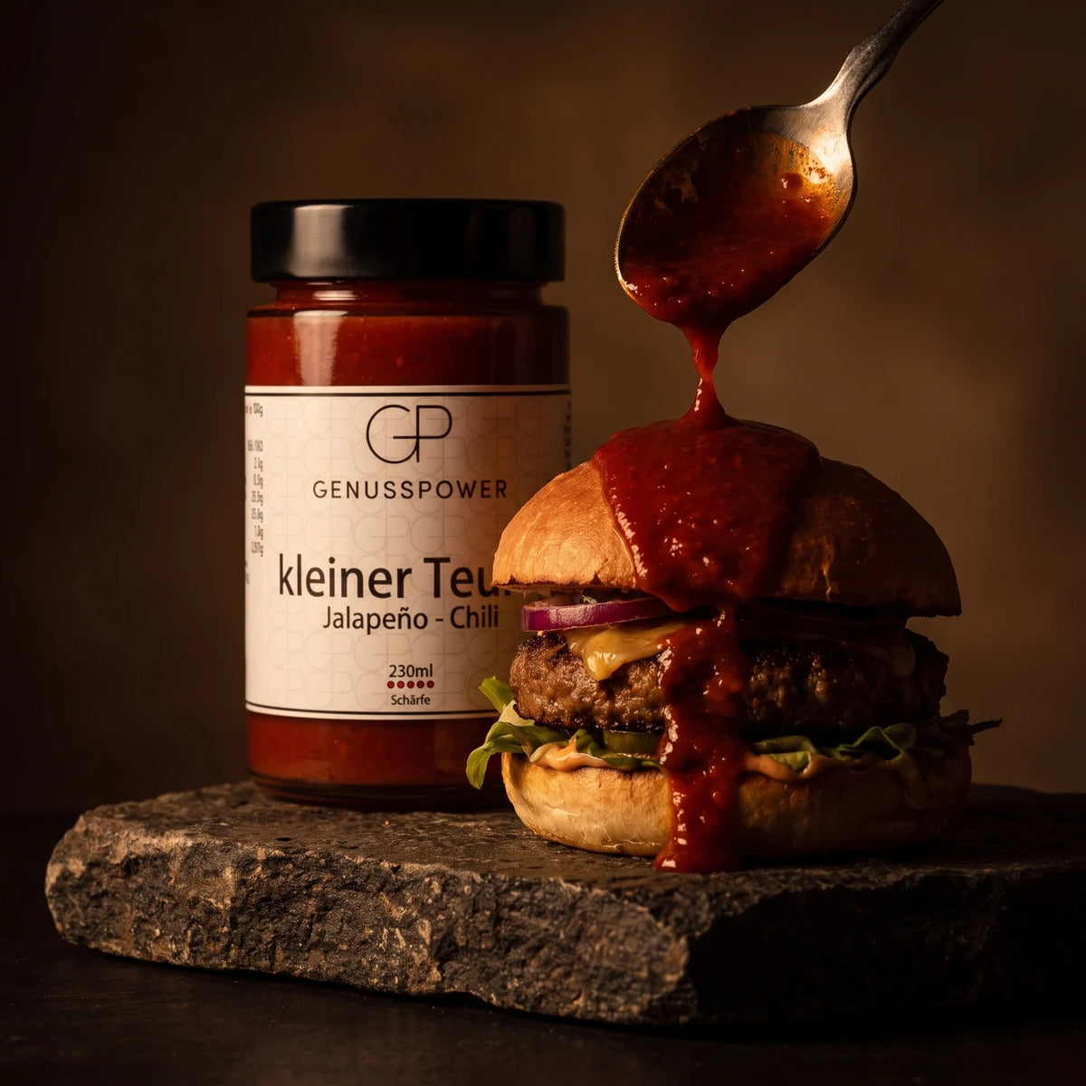 Ein Löffel gibt die Sauce auf einen Burger auf einer Steinplatte, dahinter das Glas kleiner Teufel