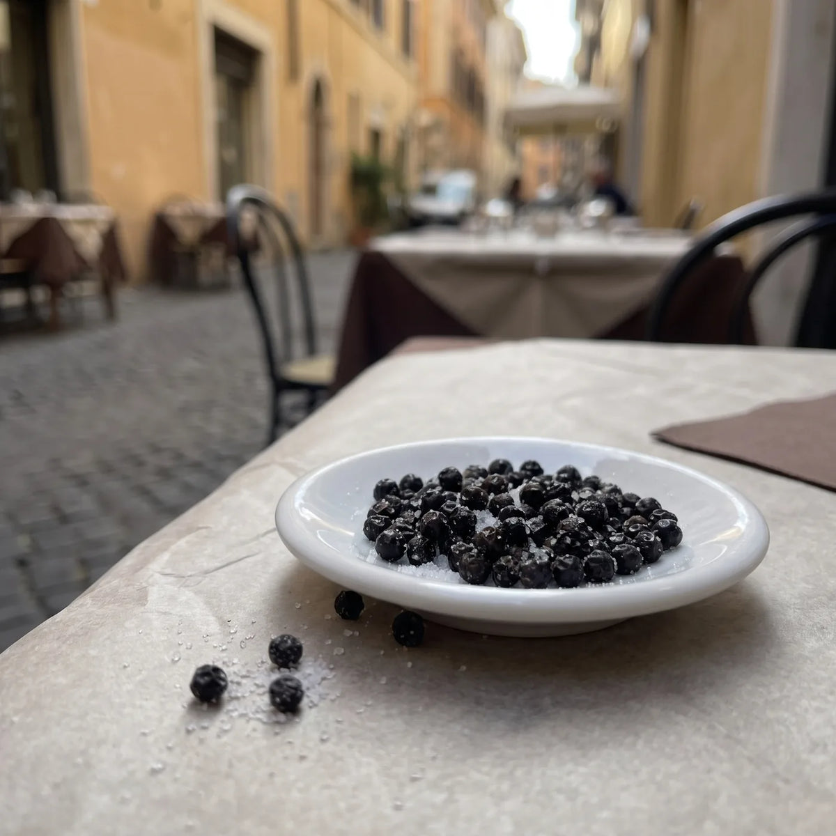 Weißes Schälchen mit glänzend schwarzen Pfefferkörnern und Salzkristallen auf einem Trattoriatisch