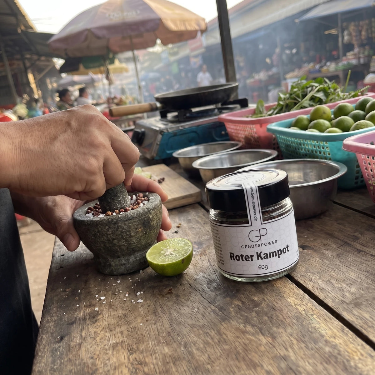 Hand zerstößt roten Kampot Pfeffer mit grobem Salz im Steinmörser an einer Garküche in Kampot, daneben das Glas und Limetten