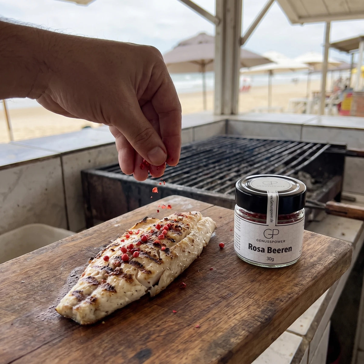 Hand lässt rosa Beeren auf ein gegrilltes Fischfilet in einer Strandbude fallen, daneben das Glas