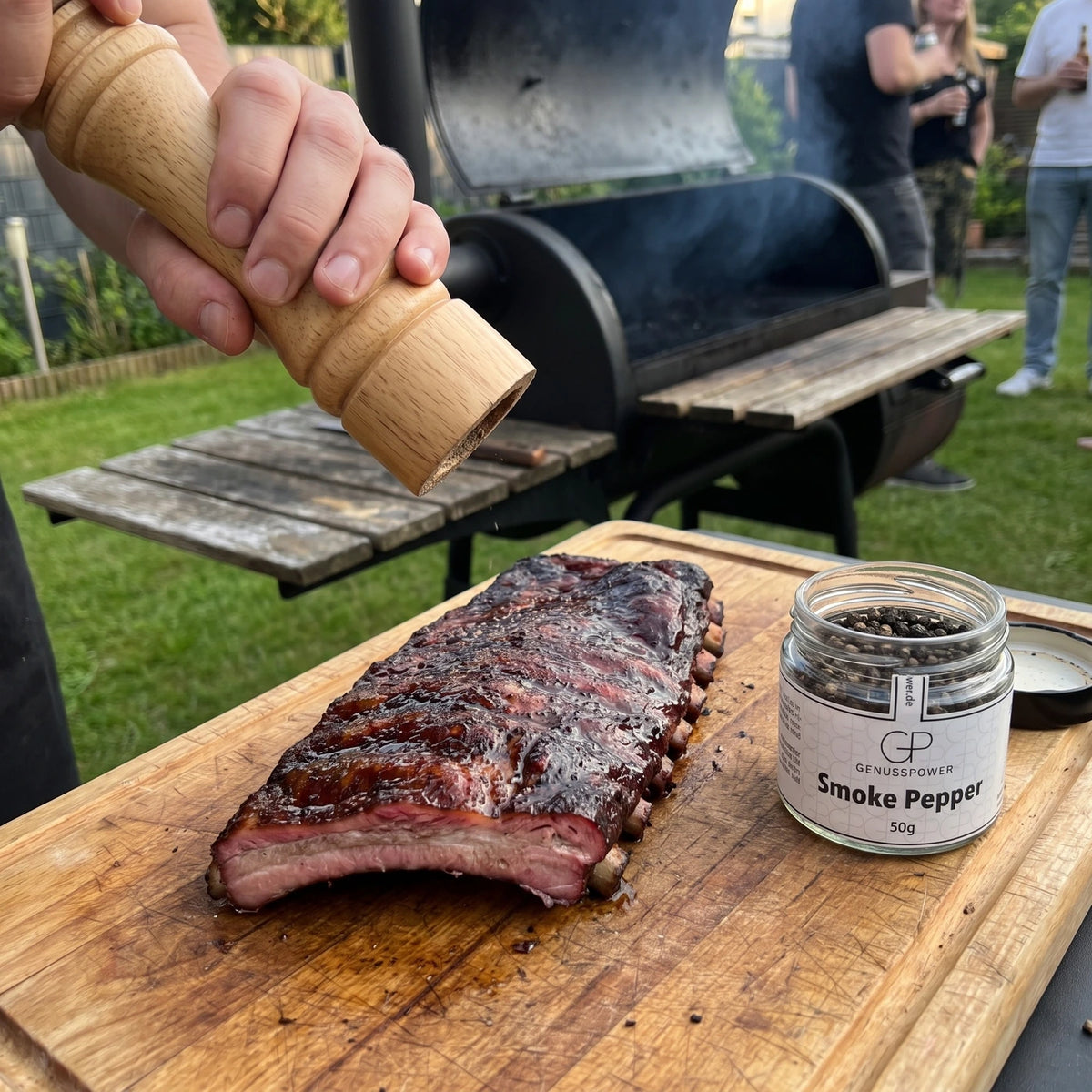 Hand mahlt mit einer Holzmühle Pfeffer über Spareribs frisch vom Smoker, daneben das Glas