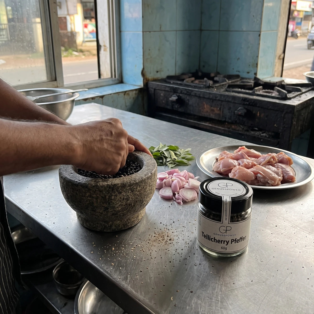 Hände zerstoßen Tellicherry Pfeffer im Granitmörser in einer Garküche in Kerala, daneben Schalotten, Curryblätter und das Glas