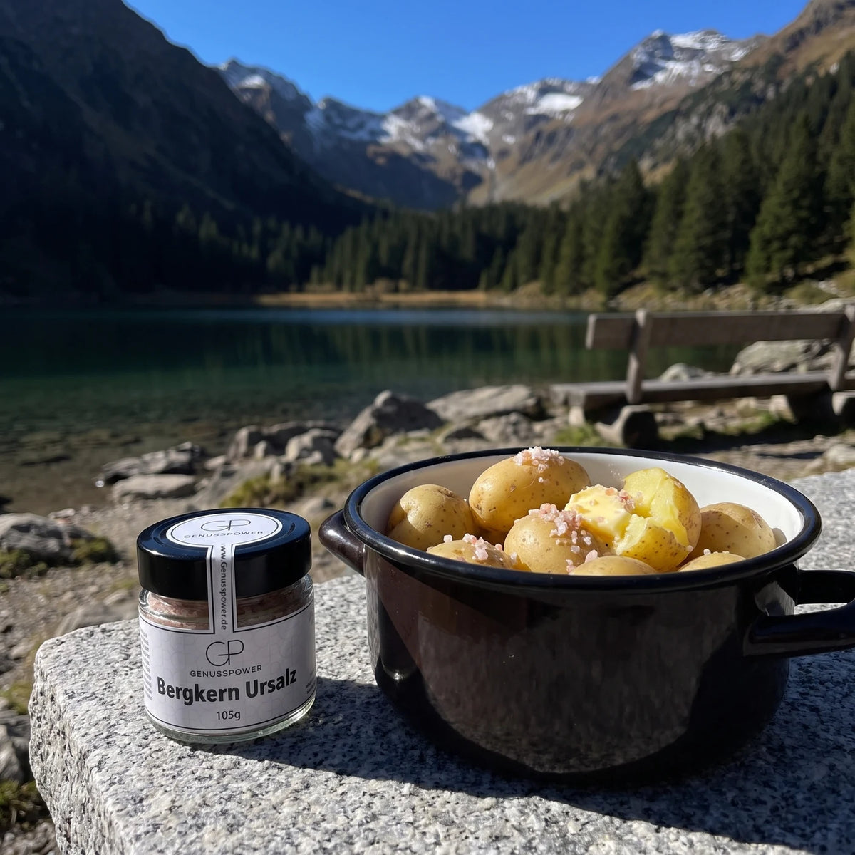 Glas Bergkern Ursalz von Genusspower auf einer Granitplatte an einem Bergsee, daneben ein Emailletopf mit Pellkartoffeln, dahinter Berge