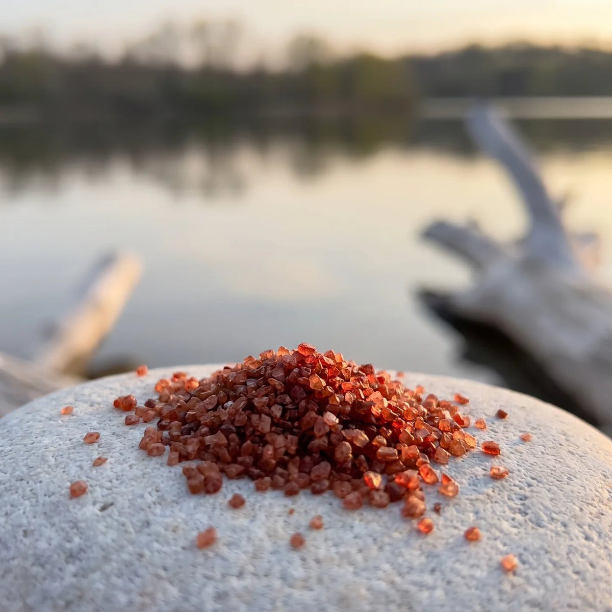 Kleiner Haufen grober ziegelroter Salzkristalle auf einem hellen Kiesel, dahinter der See im Abendlicht