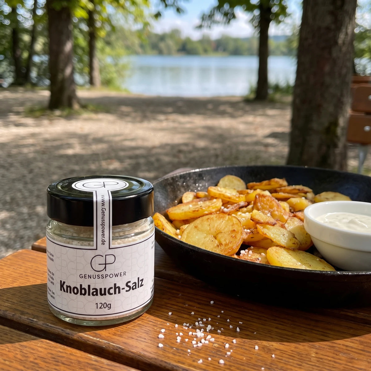 Glas Knoblauch-Salz von Genusspower auf einem Holztisch im Wirtsgarten am See neben einer Gusspfanne mit Bratkartoffeln und einer Schale Dip