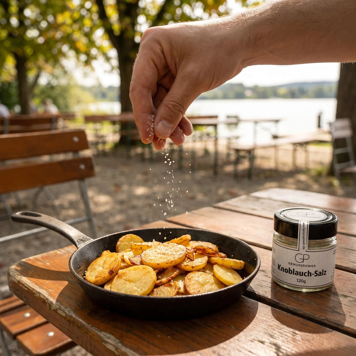 Eine Hand streut Knoblauch-Salz über Bratkartoffeln in einer Gusspfanne, daneben das Glas, dahinter Kastanien und der See