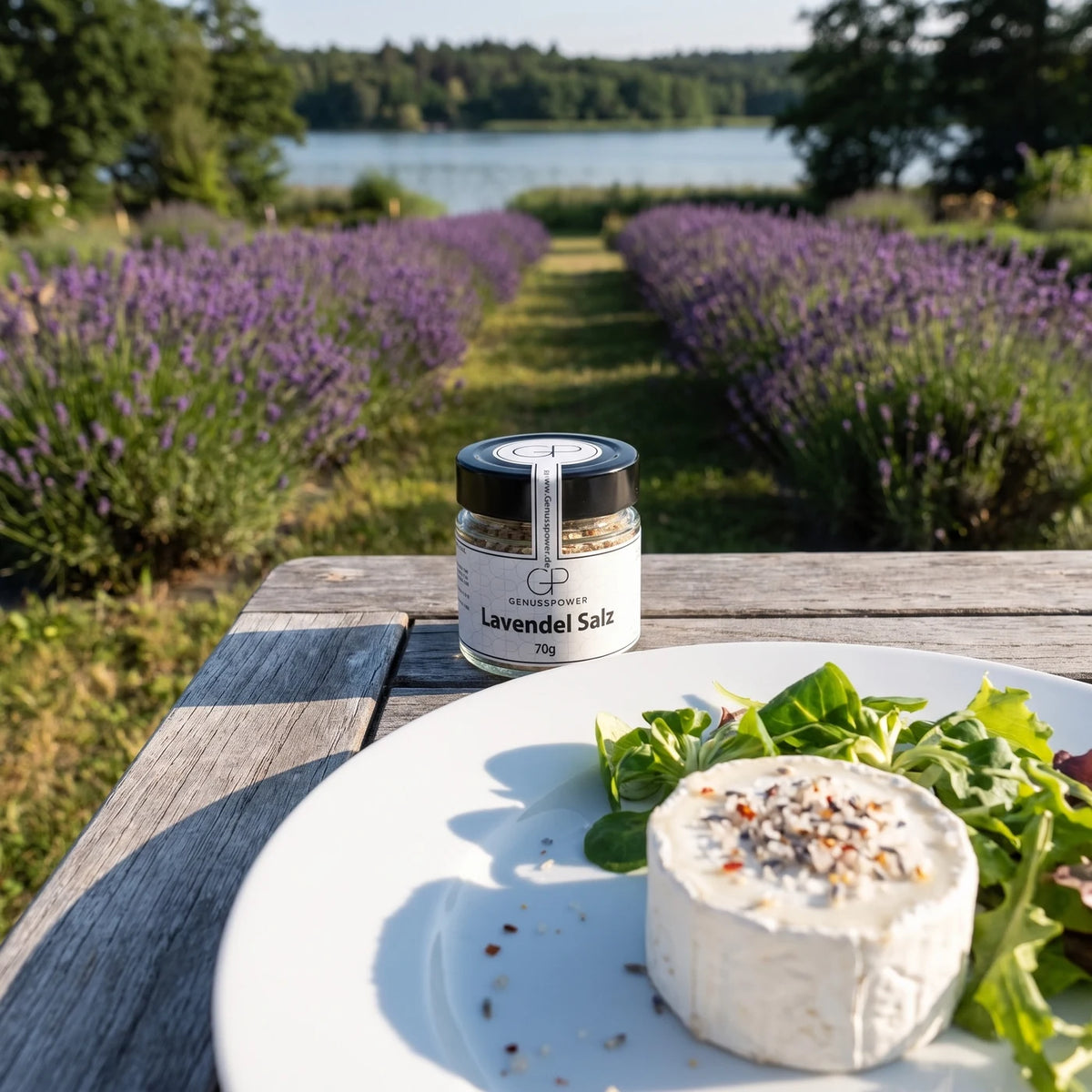 Glas Lavendel Salz von Genusspower auf einem grauen Holztisch in einem Lavendelgarten am See, davor ein Teller mit Ziegenkäse und Salat