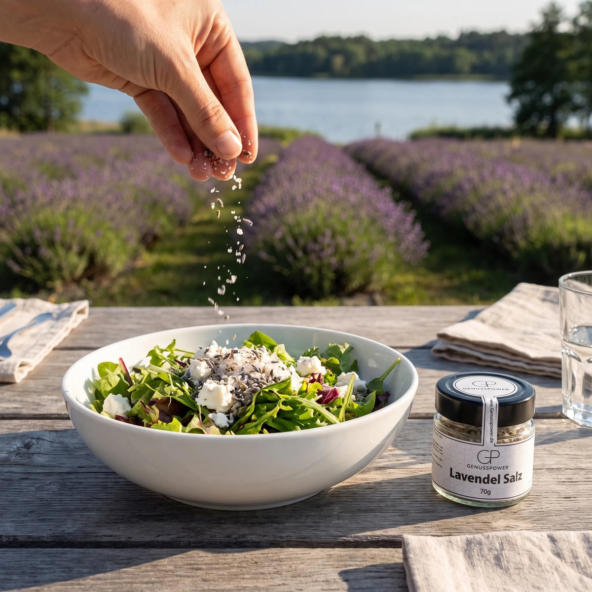 Eine Hand streut Lavendel Salz über eine Schüssel Salat mit Ziegenkäse, daneben das Glas, dahinter blühende Lavendelreihen
