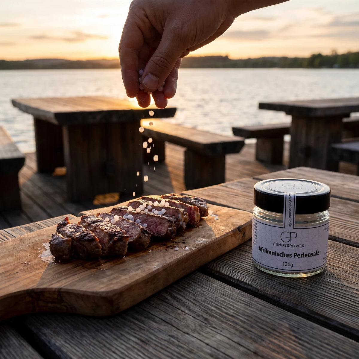 Eine Hand lässt weiße Salzperlen auf Scheiben von gegrilltem Steak fallen, daneben das Glas im Abendlicht