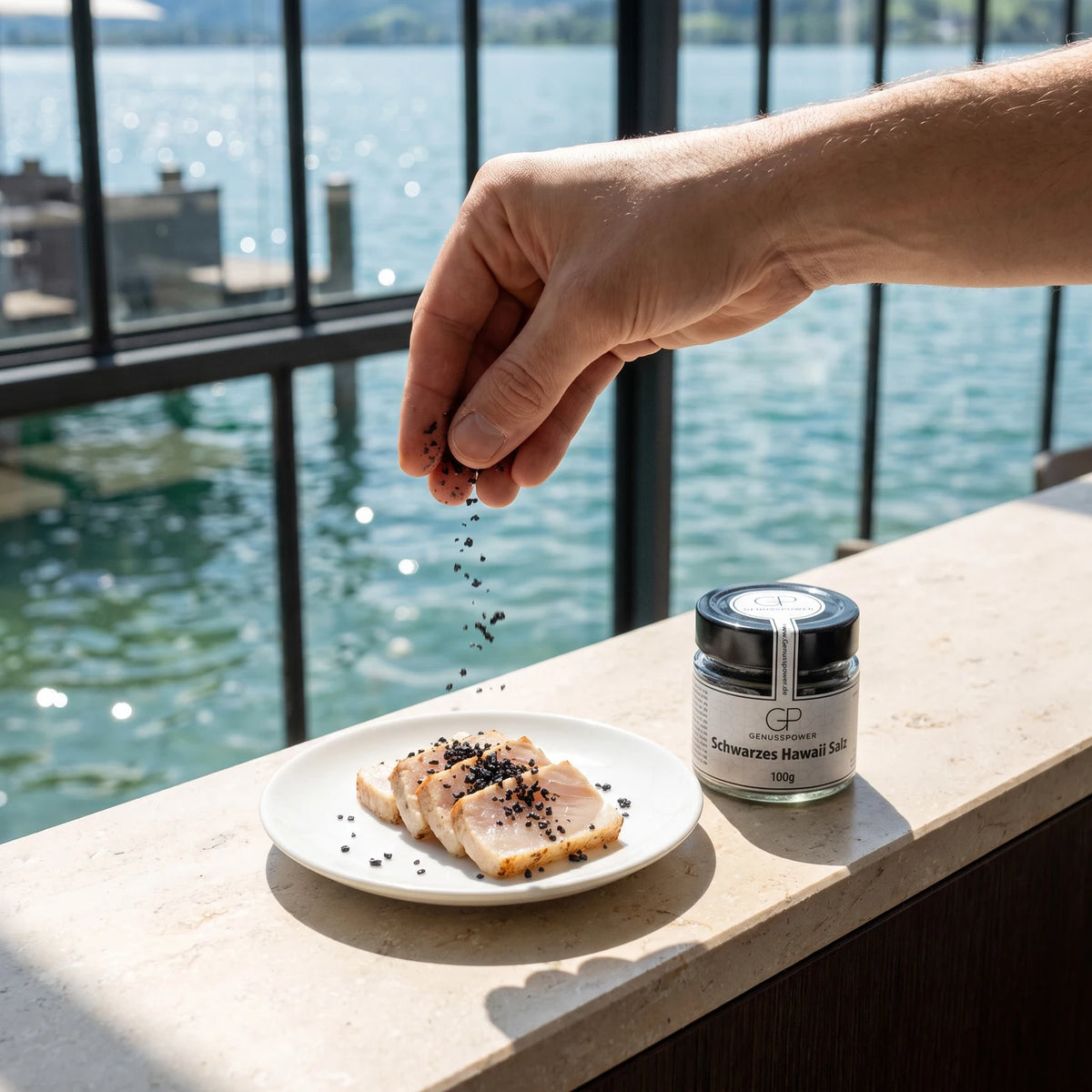 Eine Hand legt schwarze Salzkristalle auf Scheiben von hellem Fisch, daneben das Glas auf der Steintheke