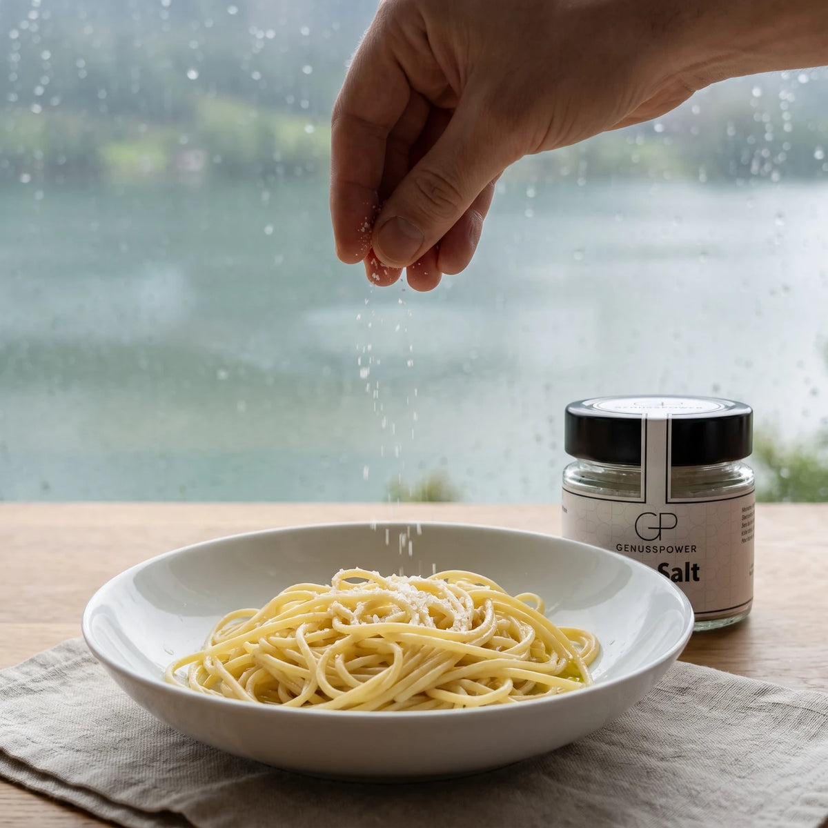 Eine Hand lässt feines Salz über eine Schüssel Spaghetti fallen, daneben das Glas, dahinter das regennasse Fenster