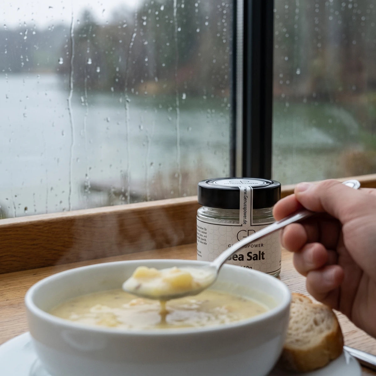 Ein Löffel hebt Kartoffelsuppe aus einer weißen Schüssel, daneben Brot, das Glas weich im Hintergrund