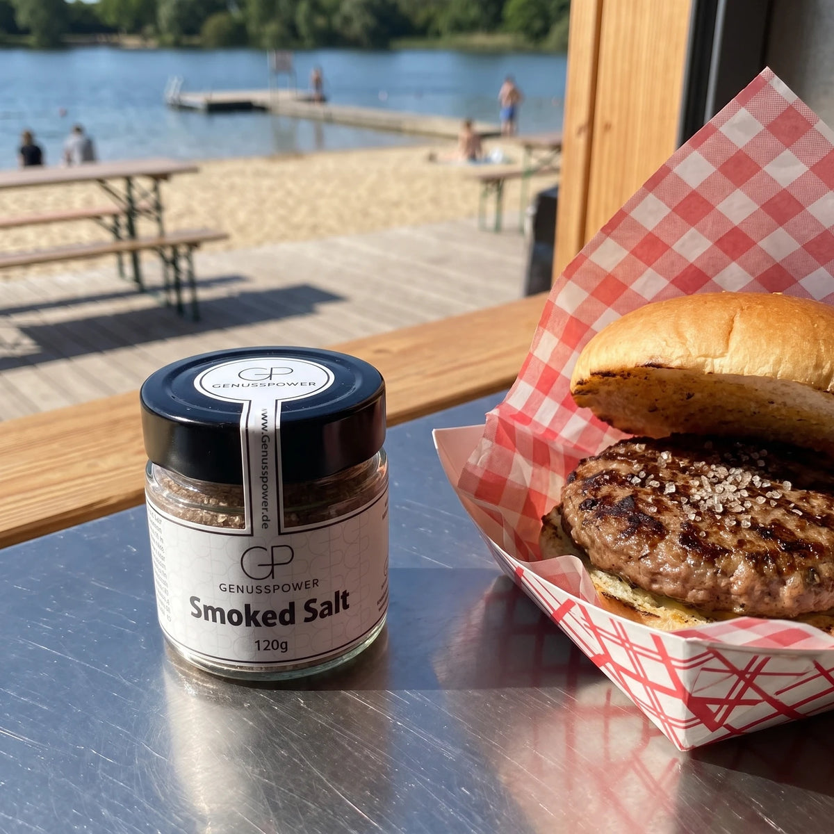 Glas Smoked Salt von Genusspower auf dem Tresen eines Kiosks am Strandbad, daneben ein Burger in kariertem Papier, dahinter Strand und See