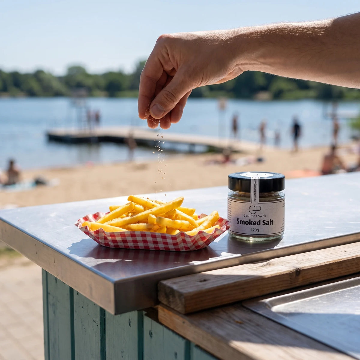 Eine Hand lässt graubraunes Salz über eine Schale Pommes fallen, daneben das Glas am Kiosk