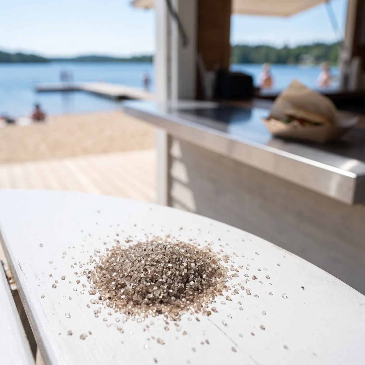 Kleiner Haufen feines graubraunes Salz auf einem weiß gestrichenen Bistrotisch, dahinter Strand und See