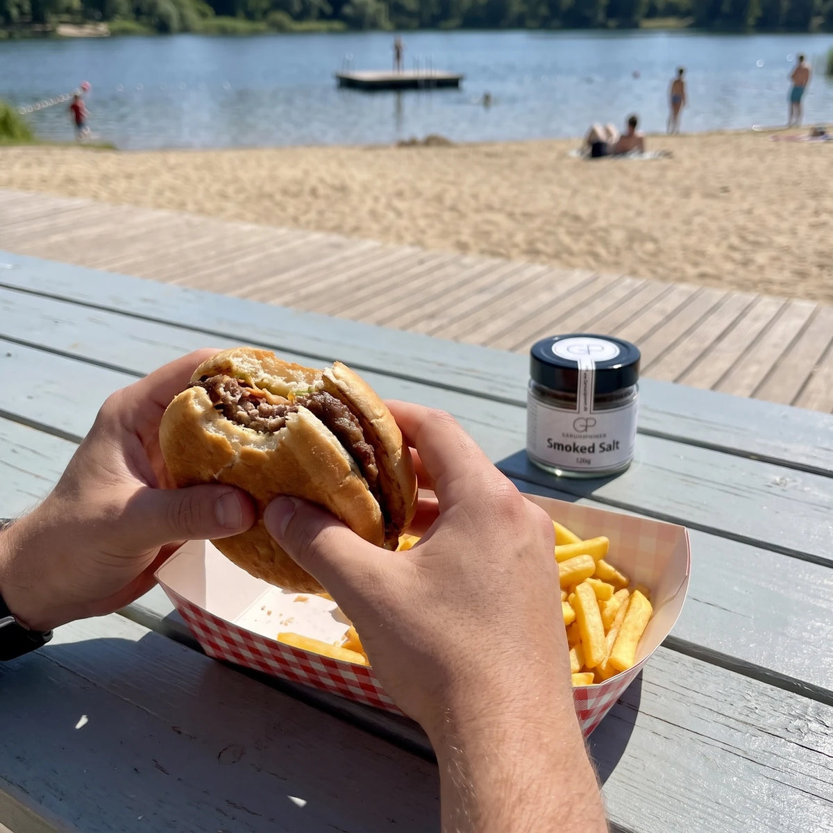 Zwei Hände halten einen angebissenen Burger, daneben Pommes, das Salzglas weich im Hintergrund