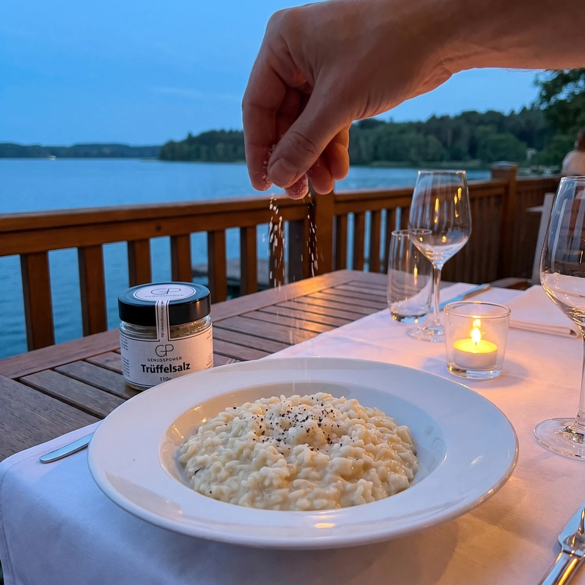 Eine Hand streut Trüffelsalz über einen Teller Risotto im Kerzenlicht, daneben das Glas, dahinter der See in der Dämmerung