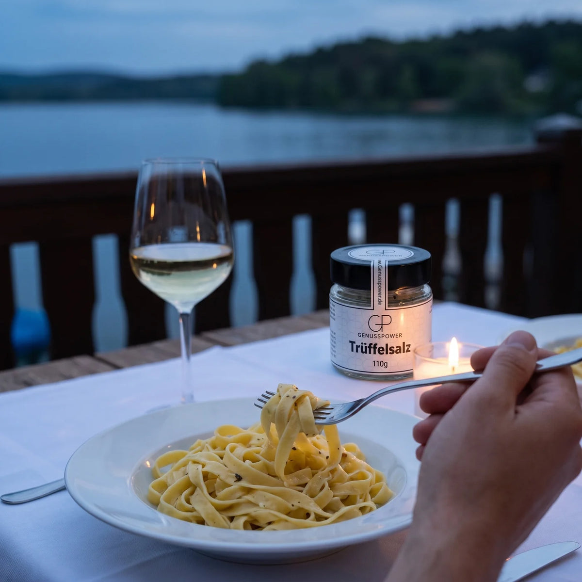 Eine Gabel dreht Tagliatelle auf einem weißen Teller, daneben ein Glas Weißwein, das Salzglas weich im Hintergrund