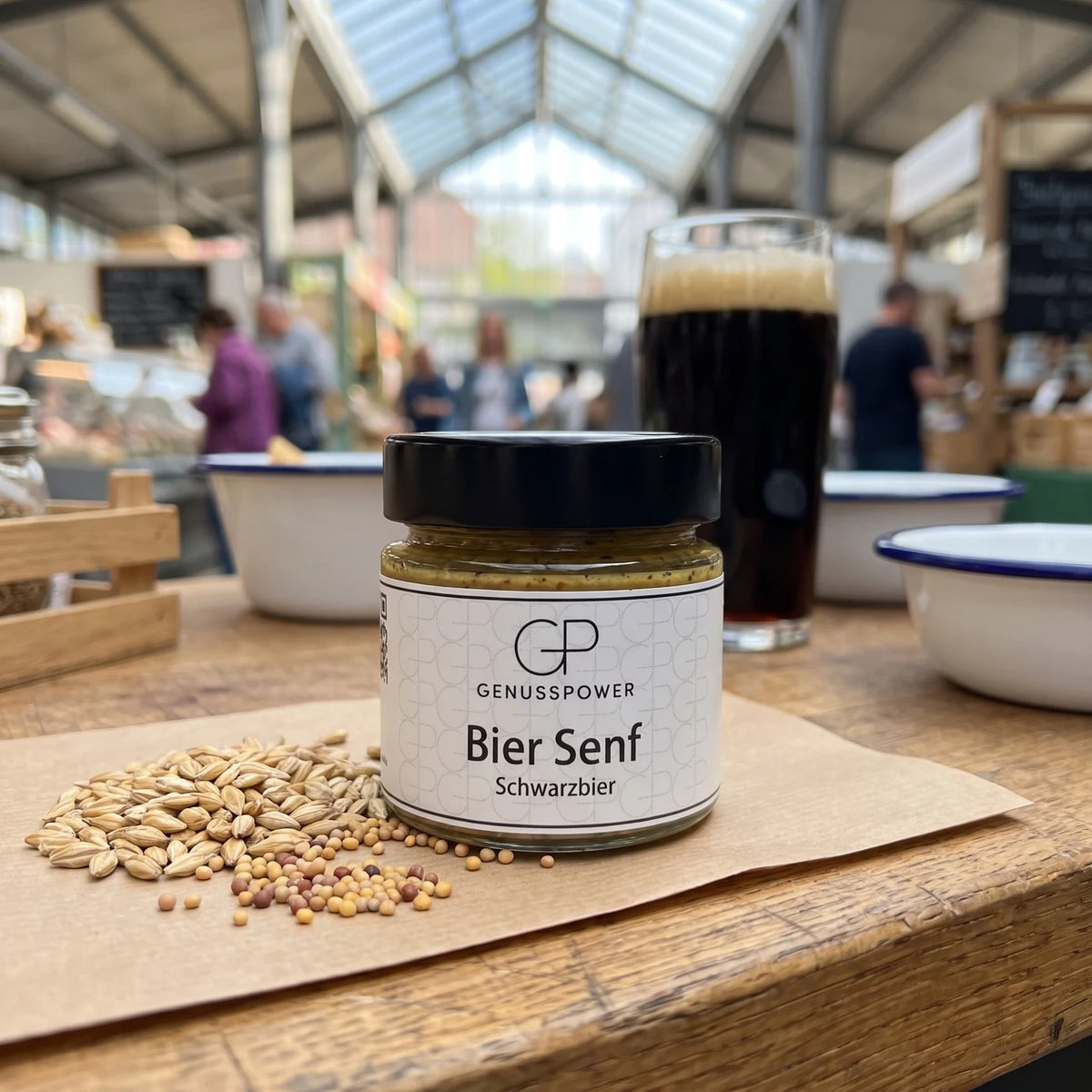 Glas Bier Senf Schwarzbier von Genusspower auf der Markttheke, daneben Malz und Senfsaat, dahinter ein dunkles Bier