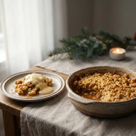 Apfel Crumble in der Auflaufform, daneben eine Portion auf dem Teller mit schmelzender Zimtschnecken Creme
