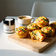 Mehrere goldgelbe Eiermuffins mit Spinat und roten Paprikastücken auf einem Brett, daneben ein Glas Rührei Eggcellent