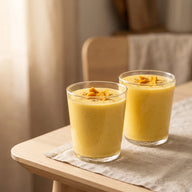 Zwei Gläser mit cremigem, goldgelbem Mango-Lassi, darüber etwas Kardamom