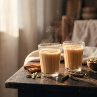 Zwei Gläser mit hellbraunem, dampfendem Milchtee, daneben Zimtstangen, Kardamomkapseln und lange Pfefferschoten