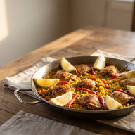 Eine große flache Pfanne mit gelbem Paellareis, gebräunten Hähnchenstücken, roter Paprika, Erbsen und Zitronenspalten