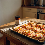 Mehrere Pizzatoasts mit geschmolzenem Käse, Schinken, Paprika und Mais auf einem Blech, daneben ein Glas Bärchen