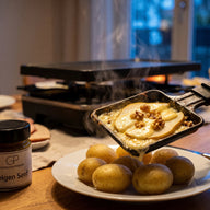 Ein Raclette-Pfännchen mit geschmolzenem Käse über Birnenspalten und Walnüssen, daneben Pellkartoffeln