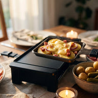 Ein Raclette-Pfännchen mit goldbraun geschmolzenem Käse über Kartoffelscheiben, Speckwürfeln und roten Zwiebelringen