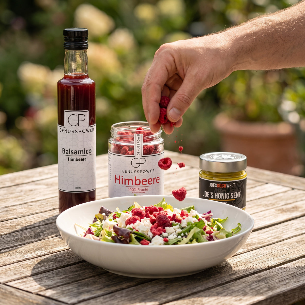 Angemacht: eine Hand streut Himbeeren aus dem offenen Himbeerglas über den Salat, daneben Balsamico Himbeere und Joes Honig Senf