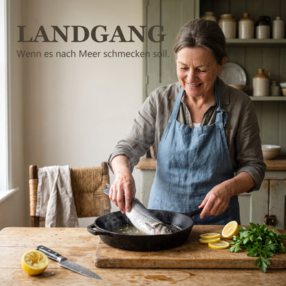 Landgang – Frau legt einen ganzen Fisch in die heiße Gusspfanne in einer Küche am Meer