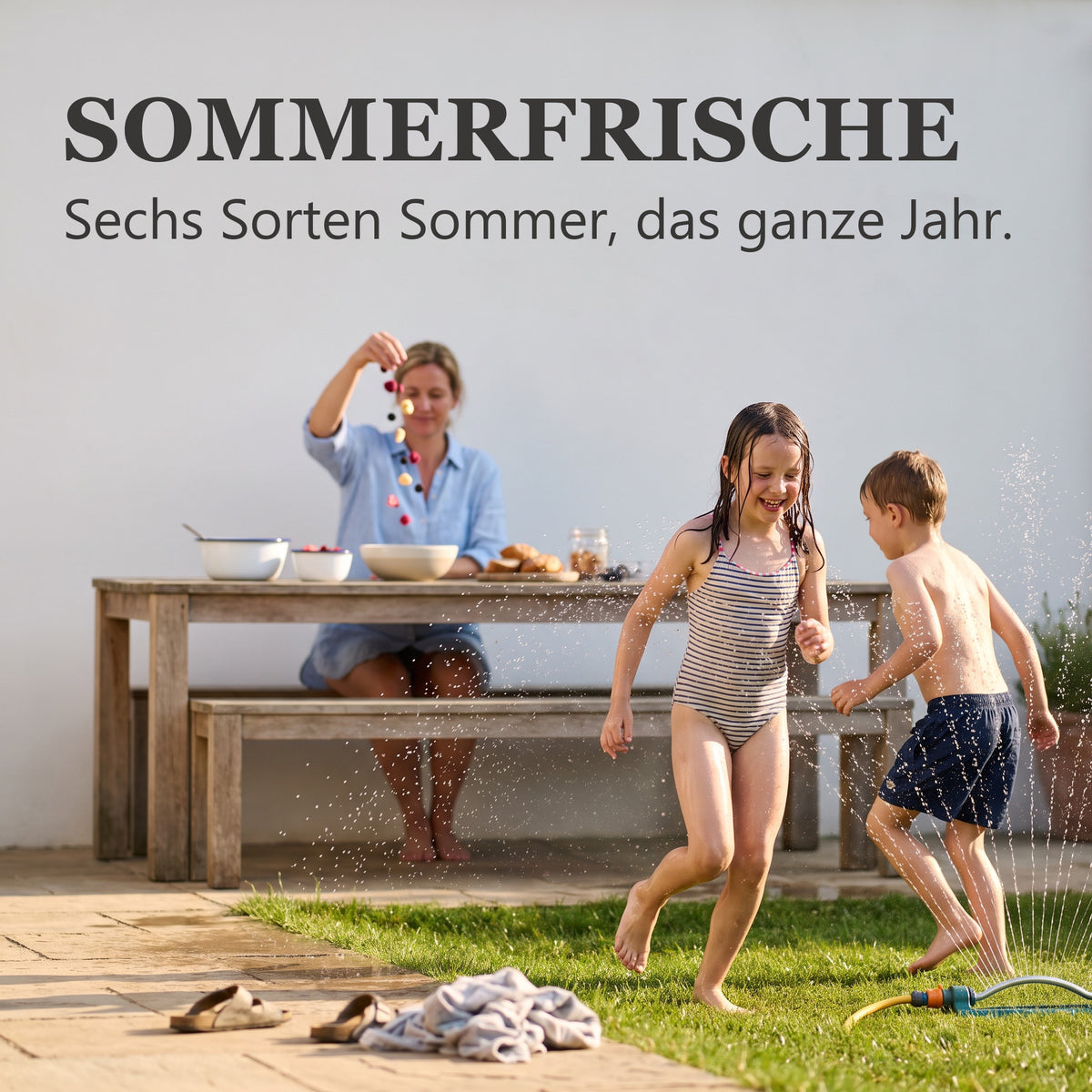 Sommerfrische: Kinder spielen im Garten am Rasensprenger, während die Mutter am Tisch eine Schale mit gefriergetrockneten Früchten anrichtet