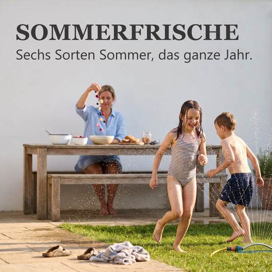 Sommerfrische – Kinder spielen im Garten am Rasensprenger, während die Mutter am Tisch eine Schale mit gefriergetrockneten Früchten anrichtet