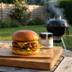 Selbstgemachter Burger mit Cheddar auf einem Holzbrett im Garten am Abend, daneben Crispy Burger von Genusspower