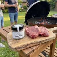 Steak Pfeffer von Genusspower neben zwei rohen Steaks auf einem Holzbrett am Grill im Garten