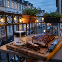 Gegrillte Spare Ribs auf einem Holzbrett auf einem Stadtbalkon am Abend, daneben Scharf auf Fleisch von Genusspower