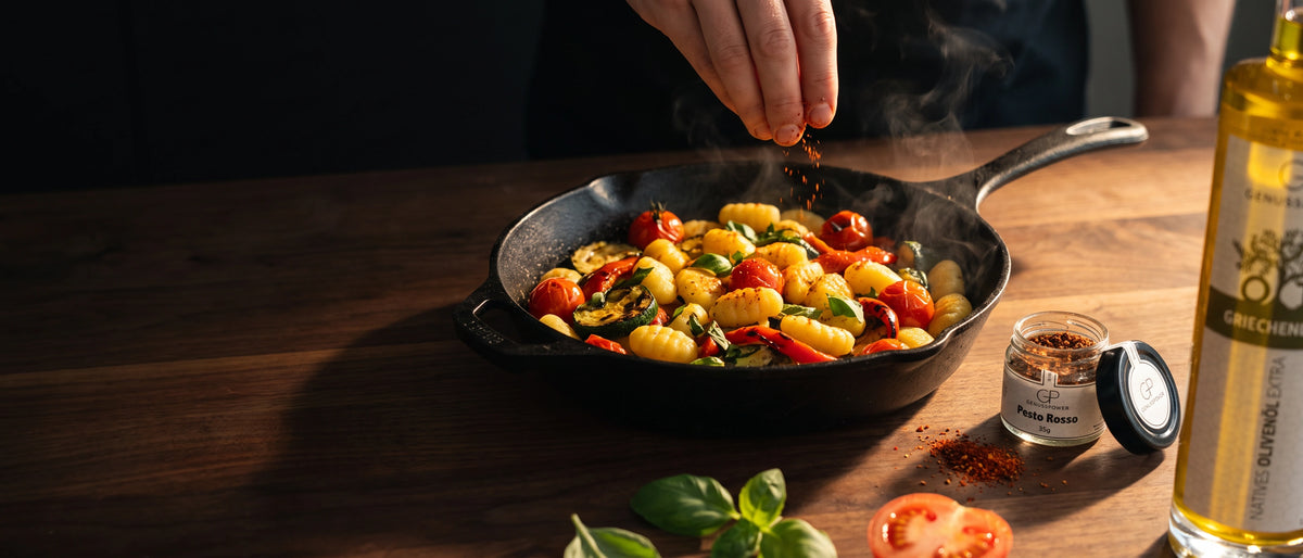 Gnocchi mit geroestetem Gemuese in der Gusseisenpfanne, eine Hand wuerzt nach, daneben Genusspower Pesto Rosso und griechisches Olivenoel