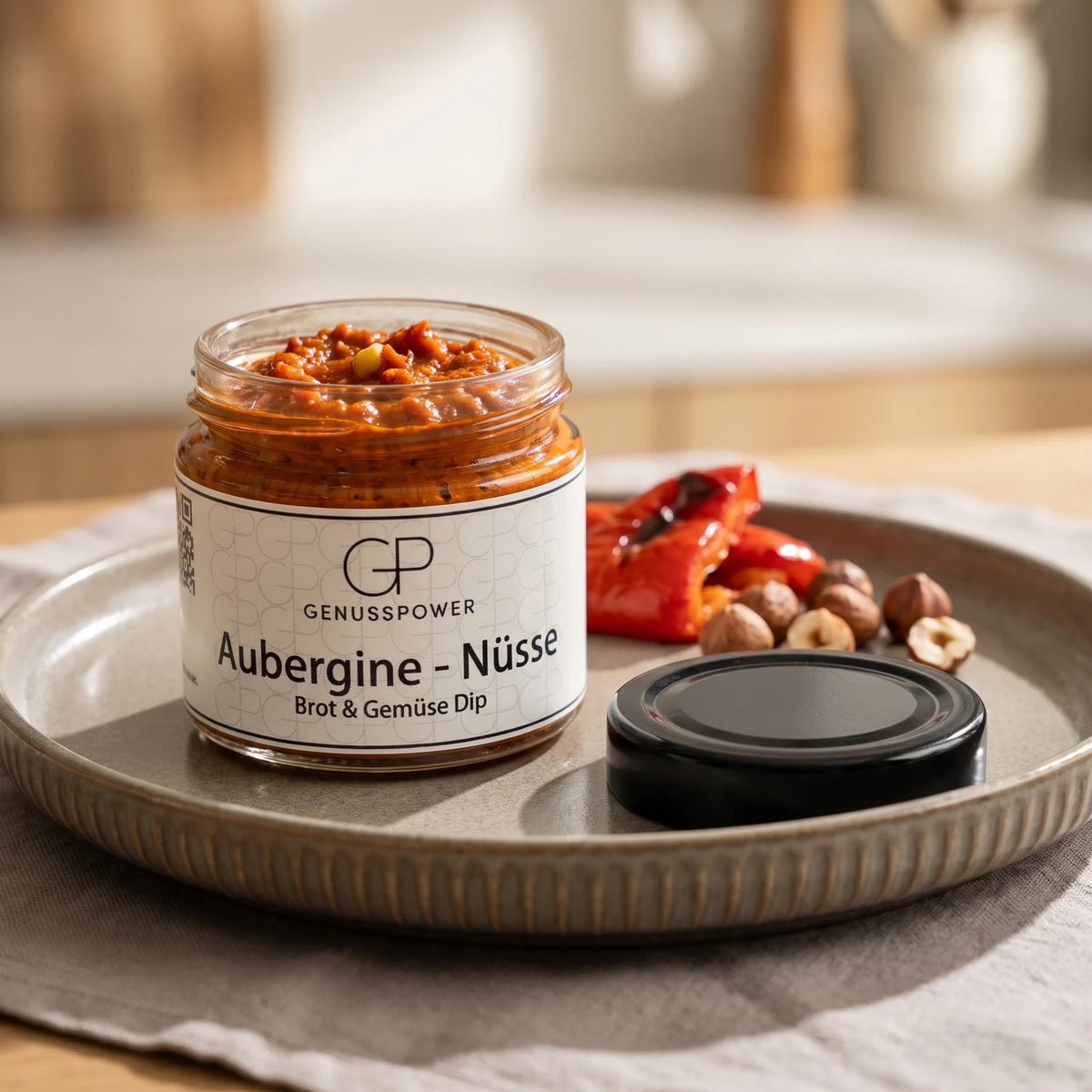 Offenes Glas Aubergine Nüsse von GenussPower auf grauer Steingutplatte, daneben eine geröstete rote Paprika und Haselnüsse